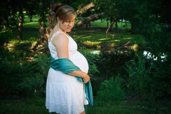 A photo of my pregnant wife, posing in front of a pond while wearing a white dress and teal scarf